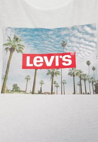 T-shirt blanc avec un imprimé graphique de palmiers sur un ciel bleu, avec un logo Levi's rouge bien en vue sur le design.