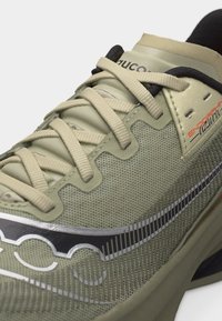 Κοντινό πλάνο ενός αθλητικού παπουτσιού σε μπεζ και πράσινο χρώμα με μπεζ κορδόνια, επάνω μέρος από δίχτυ και μαύρο-ασημί λογότυπο στο πλάι.
