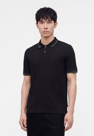 Jeune homme portant un polo noir avec des rayures multicolores sur le col et les poignets des manches, debout devant un fond blanc uni.