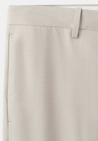 Beige-housujen vyötärönauhan lähikuva, jossa näkyy vyölenkki ja sivusauma, näyttää sileän kangaspinnan.