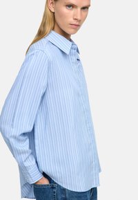Jasnoniebieska, paskowana koszula z guzikami, z kołnierzykiem w kształcie wyłogo, długimi rękawami i luźnym krojem, wykonana z gładkiego materiału o subtelnej teksturze.