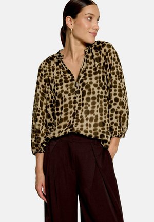 Femme aux cheveux foncés tirés en arrière, portant des boucles d’oreilles créoles dorées, un chemisier ample à imprimé léopard et un pantalon large taille haute foncé, les mains dans les poches.