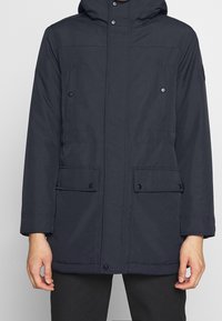 Mörk marinblå parker med huva, med dragkedja fram, tryckknappar, fyra fickor och en slät yta. Långa ärmar med manschettdetaljer.