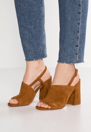 Brązowe zamszowe sandały na słupku z odkrytą palcem, paskiem wokół kostki i wycięciami. Noszone z postrzępionymi niebieskimi dżinsami.