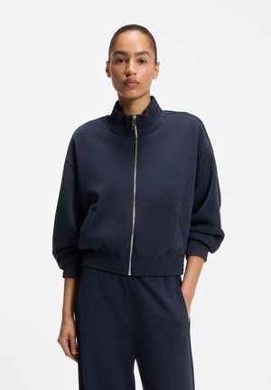 Frau steht mit einer Hand in der Tasche, trägt eine marineblaue Jacke mit Reißverschluss und passende Hose vor einem schlichten weißen Hintergrund.