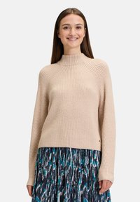 Hellbeige Rippstrickpullover mit hohem Kragen und langen Ärmeln, kombiniert mit einem plissierten Rock in Blau- und Grüntönen.