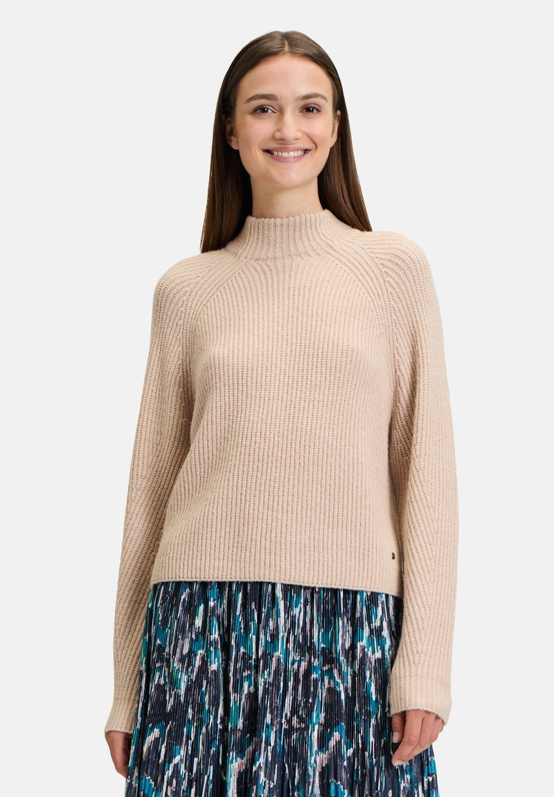 Hellbeige Rippstrickpullover mit hohem Kragen und langen Ärmeln, kombiniert mit einem plissierten Rock in Blau- und Grüntönen.