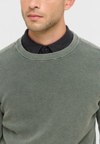 Pull en maille vert texturé avec un col rond et des bordures côtelées, superposé sur une chemise noire à col, affichant une palette de couleurs terreuses et douces.