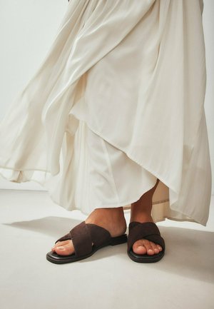 Person trägt einen fließenden weißen Rock und dunkelbraune flache Sandalen mit überkreuzten Riemen und steht auf einem hellen Boden.