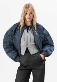 Marine bomberjakke med oversized silhuett, quiltet tekstur og glidelåsfront, lagd over en kort gray genser med kabelstrikkdetaljer.