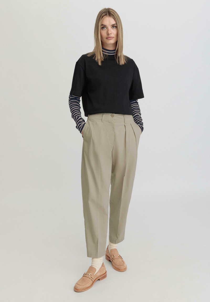 Zwart t-shirt met korte mouwen over een marineblauw en wit gestreept overhemd met lange mouwen, gecombineerd met lichtbeige geplooide broeken en bruine instappers.