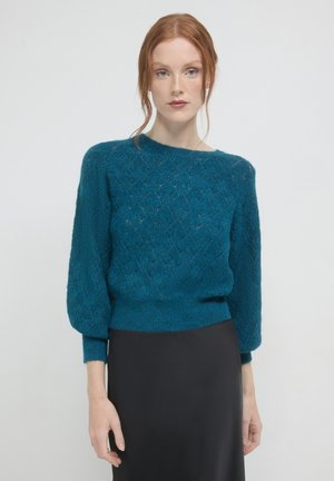 Frau mit roten Haaren, die einen strukturierten teal-farbenen Pullover mit Ballonärmeln und einen schwarzen Rock trägt, vor einem schlichten weißen Hintergrund steht.