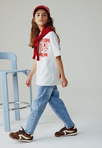 Maglietta grafica bianca con design rosso, jeans di denim blu, sciarpa rossa e sneakers marroni con accenti leopardati. Modello in movimento accanto a una sedia.