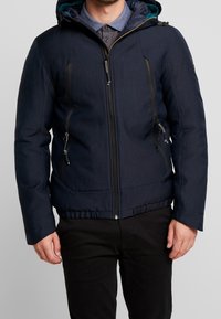 Uomo che indossa una giacca a zip con cappuccio di un blu navy scuro, con tasche a zip verticali, sopra una camicia blu con colletto e pantaloni neri.