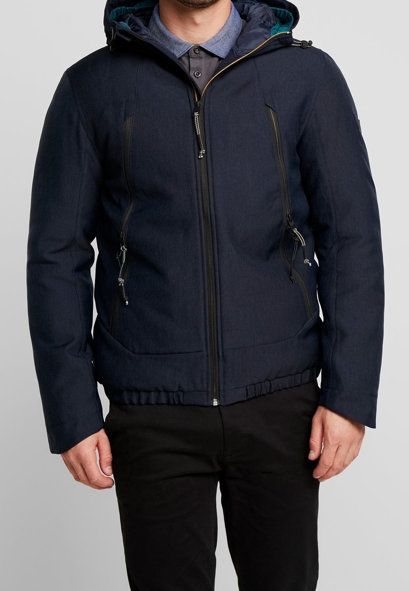 Uomo che indossa una giacca a zip con cappuccio di un blu navy scuro, con tasche a zip verticali, sopra una camicia blu con colletto e pantaloni neri.