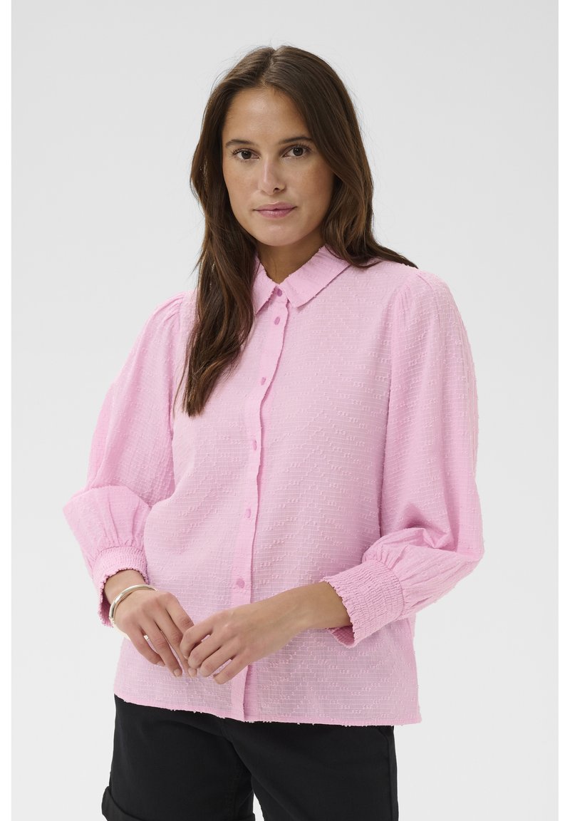 Femme aux longs cheveux bruns portant un chemisier texturé rose clair à boutons avec des manches bouffantes et un pantalon noir, debout devant un fond uni.