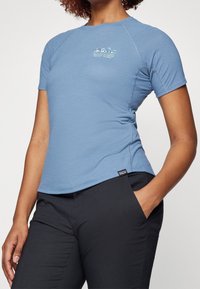 Sportovní tričko s krátkým rukávem v tyrkysově modré barvě s zakřiveným lemem a logem na hrudi. Vyrobeno z lehké látky. Kombinováno s černými kalhotami.