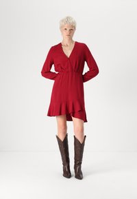 Robe rouge à manches longues, décolleté en V profond et ourlet à volants. Portée avec des bottes de cowboy en cuir marron ornées de coutures décoratives.