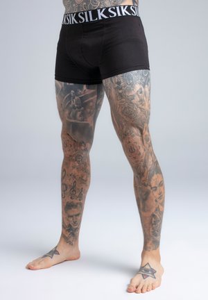 Tetovirane noge i stopala osobe koja nosi crne bokserice s pojasom "SILKS", stojeći bosi na jednobojnoj svijetloj pozadini.