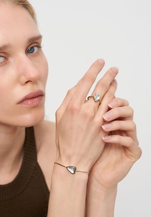 Nahaufnahme einer Person, die ihre Hand berührt, mit einem goldenen Ring und einem passenden Armband mit herzförmigen klaren Steinen.