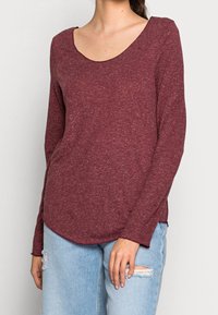 Mörkröd långärmad shirt med rundad hals, lätt böjd fåll och en texturerad tyg. Kombinerad med ljusblå slitna jeans.