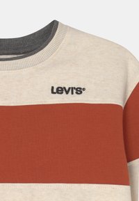 La sudadera de rayas de Levi's presenta un cuerpo beige, rayas horizontales color óxido y un cuello acanalado gris oscuro con logo bordado.