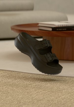 Sandale noire à enfiler avec deux boucles réglables flottant au-dessus d'un tapis beige texturé, près d'une table basse ronde en bois avec des livres empilés.