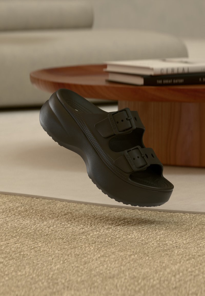 Sandale noire à enfiler avec deux boucles réglables flottant au-dessus d'un tapis beige texturé, près d'une table basse ronde en bois avec des livres empilés.