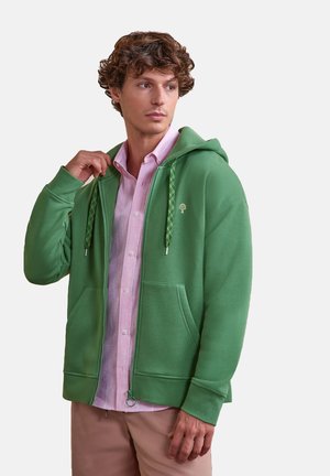 Young man wearing a green zip-up hoodie over a light pink button-up shirt, looking to the side against a plain background.