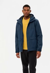 Veste bleu marine imperméable avec capuche, poches avant et cordons ajustables. Portée sur un t-shirt jaune et associée à un pantalon noir.