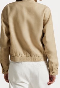 Béžová lněná bunda s nabíraným elastickým lemem, knoflíkovými manžetami a jednoduchým zadním designem. Nosit s bílými kalhotami.