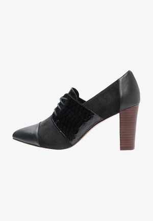 Scarpa nera con tacco, caratterizzata da una punta affilata, dettagli in camoscio e finiture lucide, un pannello in tessuto testurizzato nero e un tacco in legno impilato.