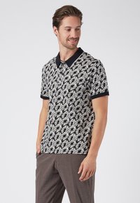 Kortärmad polo-shirt i svart och kräm med ett bladmönster, med svart krage och ärmslut, tillverkad av mjukt tyg.