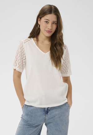 Hosszú barna hajú fiatal nő, fehér V-nyakú, csipkés ujjú felsőt és világoskék farmert visel, kezei zsebre téve, semleges háttér előtt.