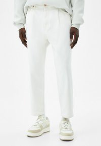 Pantalones de mezclilla blanca con un corte relajado, que presentan pliegues en el frente y detalles de costura sutiles. Combinados con zapatillas en beige y blanco.
