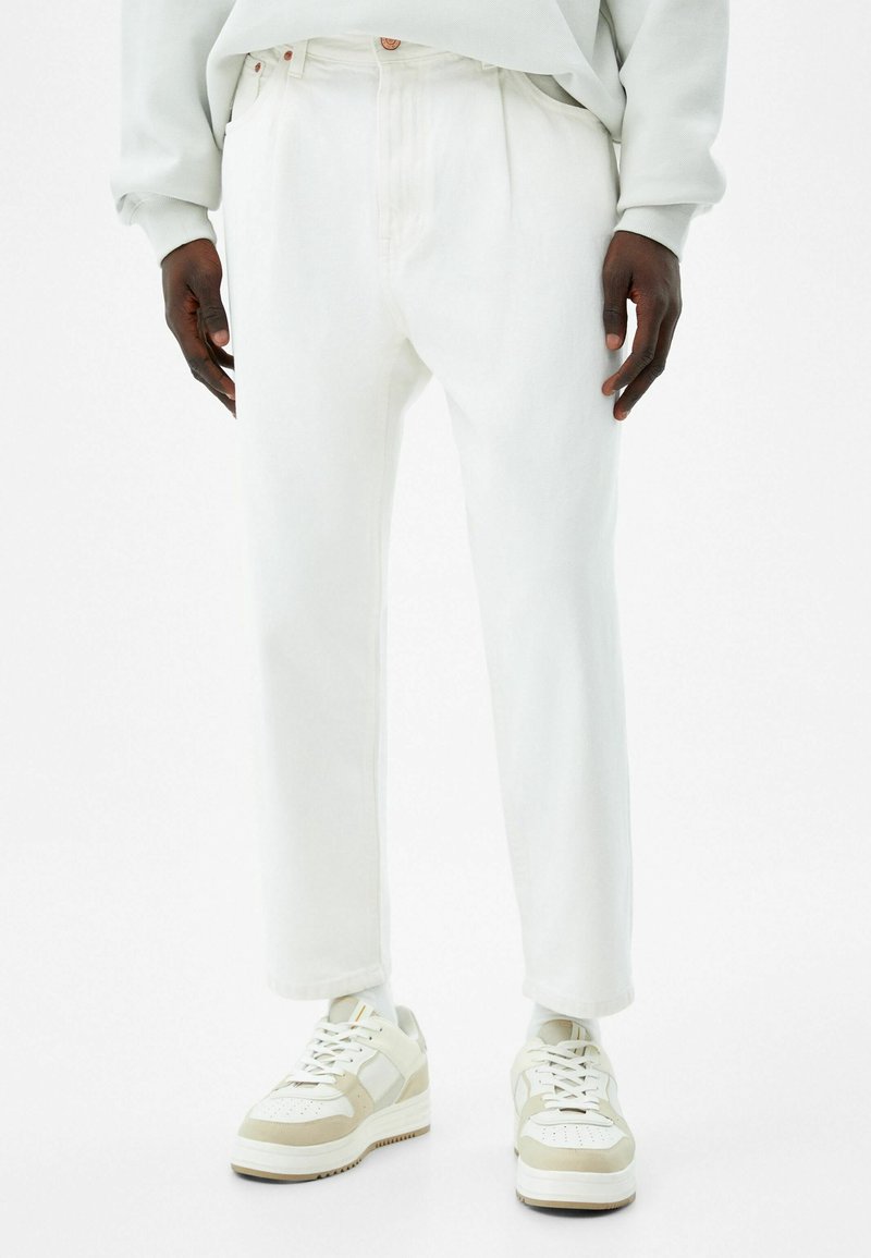 Pantalones de mezclilla blanca con un corte relajado, que presentan pliegues en el frente y detalles de costura sutiles. Combinados con zapatillas en beige y blanco.