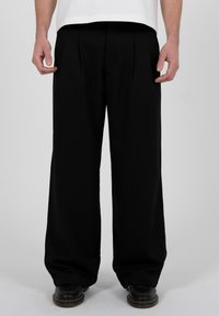 Mann, der lockere schwarze Hosen und schwarze Schuhe trägt, steht vor einem schlichten hellgrauen Hintergrund.