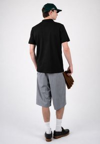 Homme de dos portant un polo noir, casquette verte, short gris à fines rayures, chaussettes blanches, chaussures noires, tenant des gants marron dans la main droite.