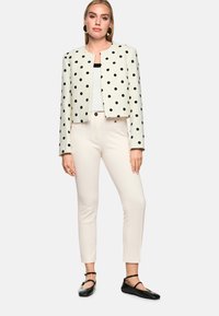 Gekürzte weiße Jacke mit schwarzen Punkten, kombiniert mit einem strukturierten schwarz-weißen Oberteil, beigen Slim-Fit-Hosen und schwarzen Ballerinas.