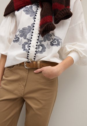 Weiße Bluse mit blauem Blumenmuster und Knöpfen, kombiniert mit beigen Hosen. Ein brauner Gürtel mit silberner Schnalle. Dunkler Schal locker über die Schultern gelegt.