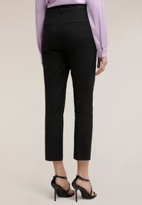 Vrouw draagt nauwsluitende zwarte broek tot op de enkels, paarse blouse en zwarte sandaaltjes met hakken, staand tegen een neutrale achtergrond.