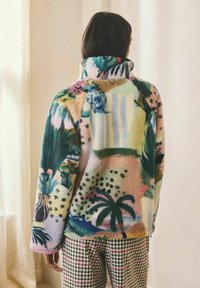 Femeie văzută de spate, purtând o jachetă fleece colorată și cu model și pantaloni în carouri, stând în interior lângă o perdea luminoasă.