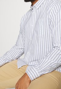 Valkoinen ja vaaleansiniraidallinen napillinen paita, jossa on tasku, kaulus ja kalvosinpituiset hihat, yhdistetty beigenvärisiin housuihin.