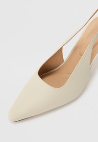 Beige slingback høyhelte sko med spiss tå, glatt syntetisk materiale, utskjærte sider og minimal sømdetalj.