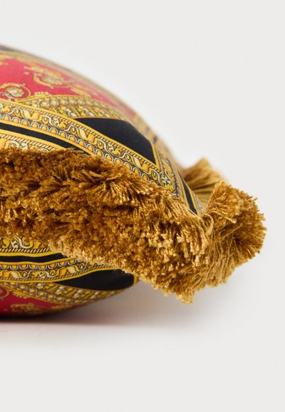 Díszpárna sarka piros, fekete és arany díszes mintával, vastag mustársárga rojttal, fehér háttér előtt.