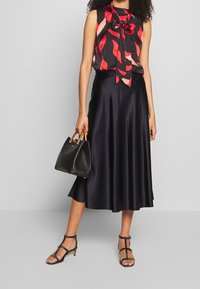 Femme portant un chemisier sans manches noir et rouge à motifs, une jupe midi en satin noire, des sandales noires, et tenant un sac à main noir avec des poignées en bois.