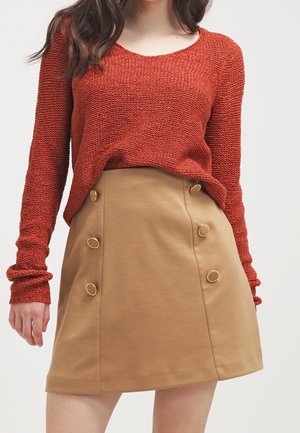 Kobieta ubrana w rdzawobrązowy dziergany sweter w połączeniu z beżową spódnicą o linii A, zdobioną dwoma pionowymi rzędami złotych guzików.