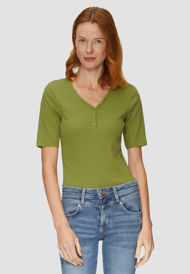 Grünes Kurzarm-Henley-Oberteil mit drei vorderen Knöpfen, im V-Ausschnitt-Design. Kombiniert mit blauen Denim-Jeans. Glatte Textur, taillierte Form.