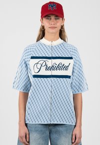 Femme portant un haut zippé bleu clair à motifs avec le texte « Prohibited » et une casquette rouge avec un logo bleu, debout sur un fond uni.