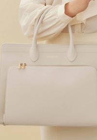 Gray leather tote bag with top handles, featuring a front zip pocket adorned with gold hardware and brand logo. Smooth, textured finish.
